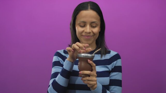 Woman holding mate cup over pink background seems thoughtful in striped sweater looking sideways on an isolated bright wall adding an intriguing atmosphere to the image.