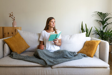 Woman reading at home drinking cup of coffee looking away. Female relaxing at home reading book enjoying herbal tea.