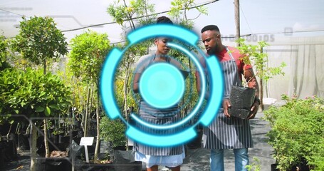 Holding black pot with sapling, two workers in striped aprons consulting notepad in nursery aisle
