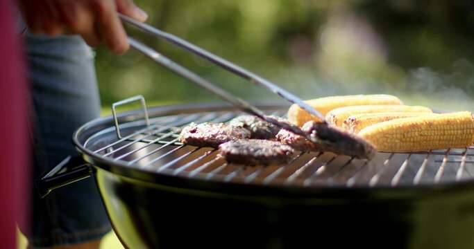 Grilling burger patties and corn on charcoal kettle grill in backyard, wearing denim and red top - Powered by Adobe