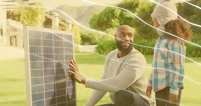 Holding solar panel, kneeling father talking to daughter in front yard, with knit hat and sweater