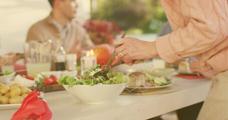 Tossing adult in pink cable-knit sweater using tongs, serving salad from white bowl at yard-table
