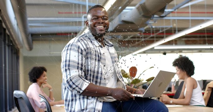 Smiling man wearing plaid shirt sitting on long wooden table holding laptop, working in open office