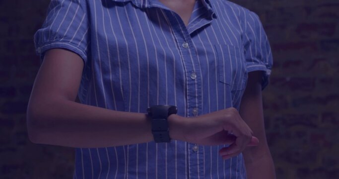 Pointing, woman adjusting smartwatch on left wrist at brick wall in blue shirt under blue light