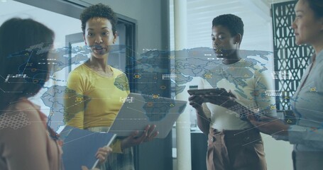 Presenting four women in office reviewing data with map overlays, yellow top holding laptop tablet