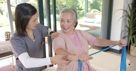 Stretching Asian woman in pink pulling blue band on table at home, therapist grey scrubs guiding
