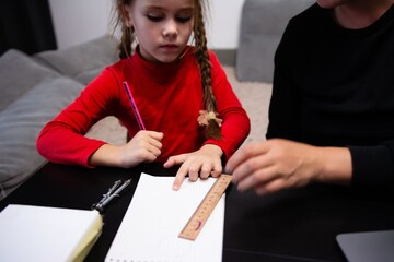 A child in a red shirt is doing homework at a table with a ruler, pencil, compass and notepad. The teacher is assisting the child.