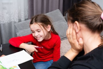 A smiling young girl in a red shirt sits at a table drawing, while a woman sits behind her with her...