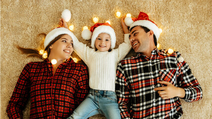 Top view of a Joyful family lying on the carpet with glowing lights, mother and father in Santa hats smiling at their daughter in the middle, celebrating love, Christmas eve festive atmosphere.