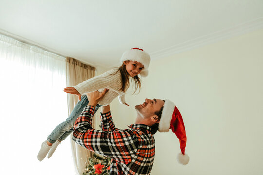 Happy father lifting his daughter in the air while both wear Santa hats, smiling joyfully and playing together during the Christmas holiday season