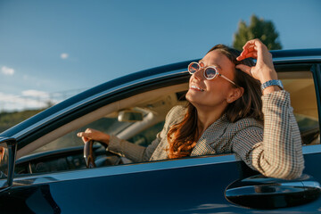 sunlit woman leaning from car window smiling, sunglasses lifted, enjoying golden-hour drive on scenic road, relaxed sweater,