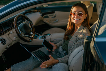 Woman closing business deal from car, smiling confidently while juggling laptop and phone, executive energy and poised body