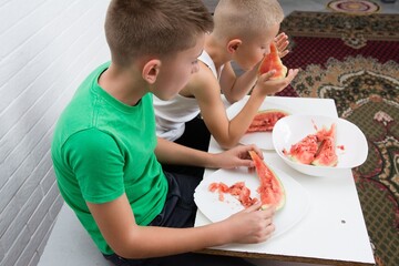 Two boys enjoying watermelon indoors. Eating the fruit at a table. Candid and natural. Summer treat. Childhood moment.