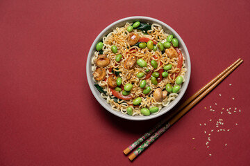 Asian noodle bowl with edamame beans, mushrooms and vegetables on dark red background, minimalist top view of healthy vegan meal
