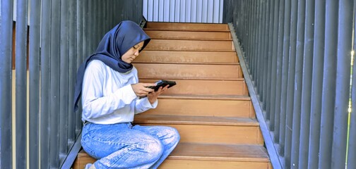 woman wearing hijab sitting on wooden stairs holding a digital tablet