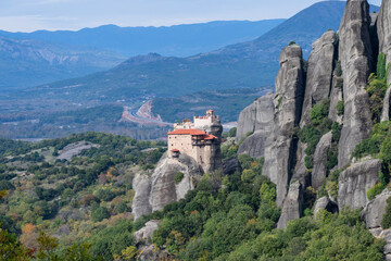 A monastery with red-tiled roofs crowns a steep rock pillar in Meteora, surrounded by lush forests and dramatic cliffs.
