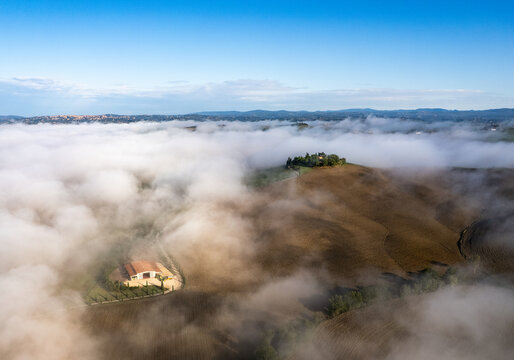 Aerial view of a serene, ethereal landscape, where the rolling hills peek through a soft blanket of fog, casting shadows and highlighting the golden hues of the fields, Val dâ€™Orcia, Tuscany, Italy.