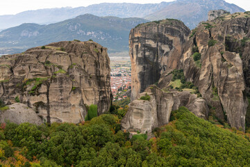 Towering rock cliffs rise from a forested valley, framing a distant town beneath a cloudy mountain sky.