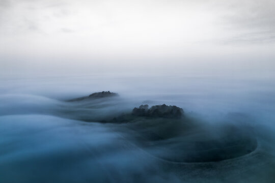 Aerial view of the Wittenham Clumps shrouded in dense fog, a mystical landscape of muted blues and grays, Oxfordshire, England, United Kingdom.