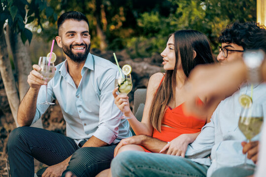 Diverse friends socializing with cocktails at an outdoor summer party. Happy multi-ethnic group of young people talking, smiling and drinking together, enjoying a moment of friendship and leisure.