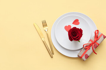 Beautiful table setting with red rose, paper hearts and gift box on yellow background. Valentine's Day celebration