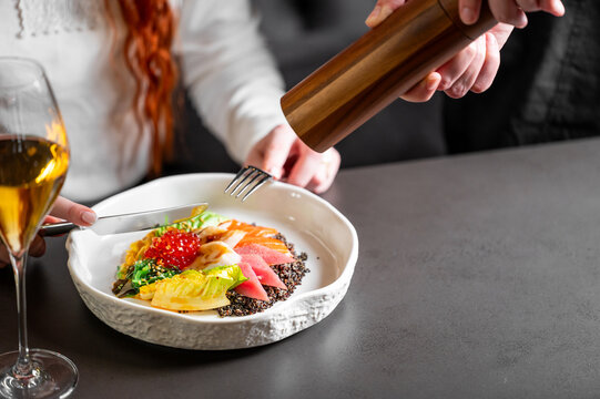 A close-up shot of hands seasoning a healthy, gourmet poke bowl or salad with a wooden pepper grinder at a restaurant table with a glass of white wine