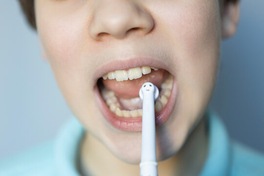 Child brushes teeth with a toothbrush featuring a smiley face while wearing a blue shirt in a well-lit room