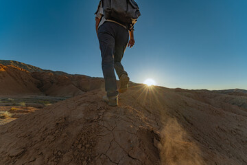 A lone traveler walks up a sandy hill in the Mongolian desert at sunset during golden hour. The sun dips below the horizon, highlighting the boot sole in the foreground.