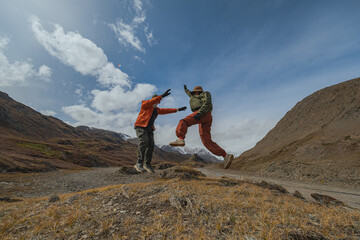 A joyful couple in bright clothing jumps playfully against a backdrop of autumn mountains, frozen mid-air in funny poses. A snow-dusted valley stretches behind them.