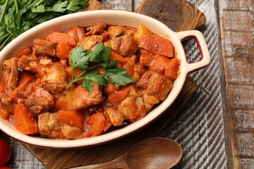 Delicious cooked stew in baking dish with fresh parsley and spoon on wooden table, flat lay