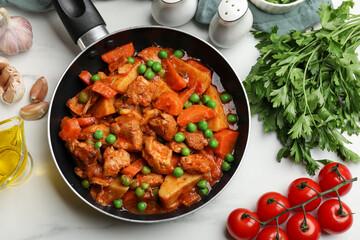 Delicious cooked stew in frying pan and ingredients on white marble table, flat lay