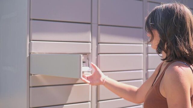 woman accesses mailbox in sunlit courtyard, casual woman retrieves mail from steel locker outdoors, outdoor setting with woman opening parcel locker to collect mail in natural light