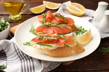 Tasty bruschettas with salmon, cream cheese, cucumber, arugula and lemon on wooden table, closeup