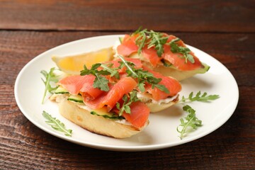Tasty bruschettas with salmon, cream cheese, cucumber, arugula and lemon on wooden table, closeup