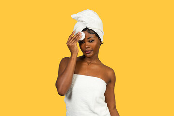 Black woman wrapped in towel using cotton pad for skincare