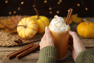 Woman holding tasty pumpkin latte with whipped cream at wooden table against blurred lights, closeup. Bokeh effect