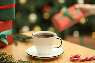 Woman holding gift box and wooden table with aromatic coffee, candy cane and fir tree branches against blurred lights, selective focus. Christmas greeting card
