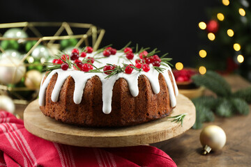 Delicious Christmas cake with icing, cranberries and rosemary on wooden table against black background with blurred lights, closeup. Bokeh effect