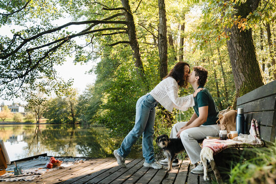 Couple kissing on lakeside bench with dog and picnic outdoors