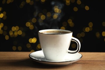 Aromatic coffee in white cup on wooden table against black background with blurred lights, closeup. Bokeh effect