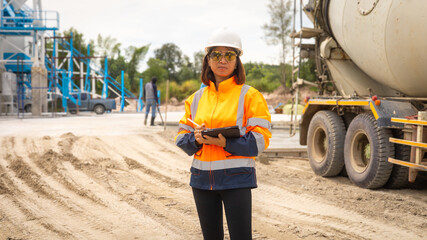 A construction site supervisor wearing safety gear stands on a sandy area, holding a clipboard while overseeing operations. Workers can be seen in the background operating machinery