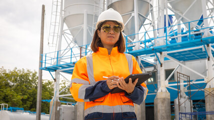 A construction site manager wearing an orange safety jacket and hard hat is taking notes on a tablet. She stands in front of silos, monitoring operations in bright daylight