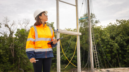 A construction worker, wearing a safety jacket and helmet, examines plans while observing power...