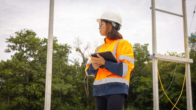 A construction worker wearing a bright safety jacket and hard hat stands on-site, holding a tablet and observing ongoing work among trees and equipment during daylight