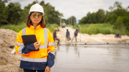 A woman in a hard hat and safety jacket stands by a waterway, holding a clipboard. In the background, workers are seen engaging in construction activities. It is daylight and the site looks busy © Happy Photo