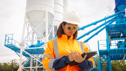 Fototapeta premium A construction site worker in a bright orange safety jacket and hard hat is checking notes on a clipboard. Large concrete silos are in the background, indicating ongoing construction work