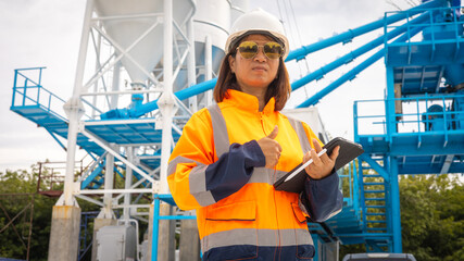 Fototapeta premium A construction site manager stands confidently at an industrial location wearing safety gear. She holds a tablet and oversees operations while surrounded by machinery and equipment