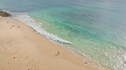 Tropical beach in Bali featuring people enjoying the sand and clear turquoise water, with surfers waiting for waves and swimmers splashing at the shoreline, captured from an aerial perspective