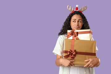 Sad young African-American woman in pajamas and reindeer horns with gift boxes on lilac background....
