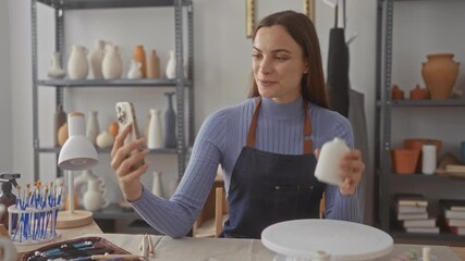 Woman holds ceramic vase and smartphone, hand visible while inspecting pottery in studio; creativity focus.
